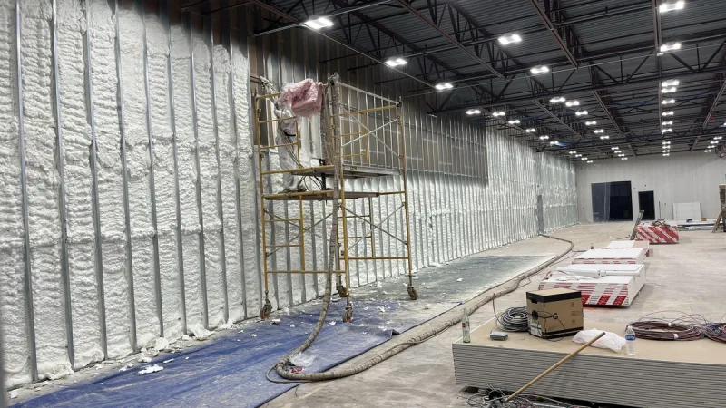 Spray foam applied to the walls of a commercial warehouse under construction, rolling scaffold and hose run on the slab below open-web steel joists