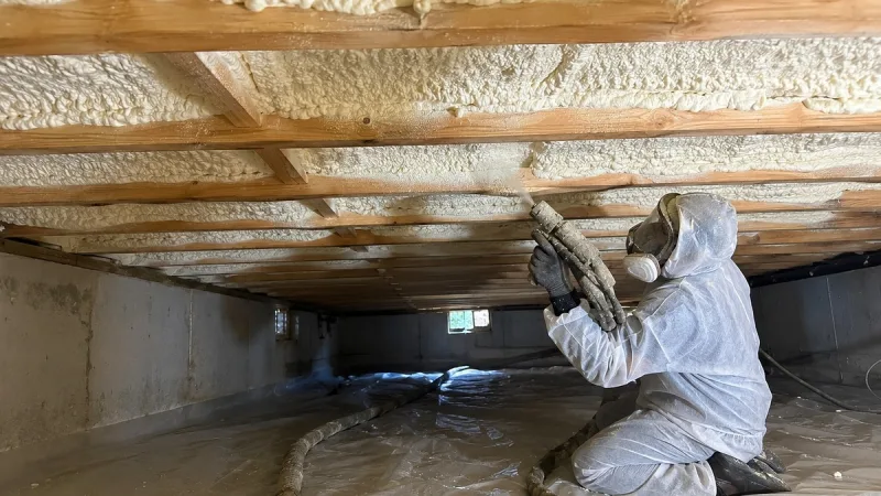 Technician in a respirator spraying foam onto the underside of the floor in a crawl space, over a ground vapor barrier