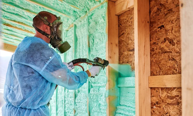 Certified applicator in a full-face respirator and coveralls spraying foam into a wall cavity
