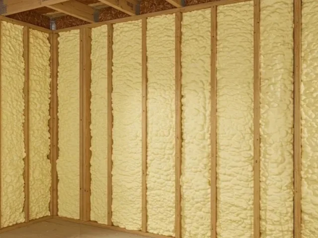 Open-cell spray foam filling every stud bay of a framed wall, pale yellow and packed flush with the stud faces